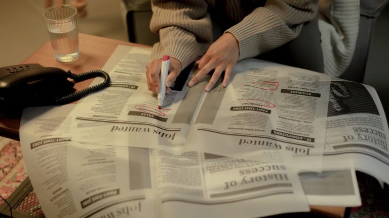 An adult woman marks job listings in a newspaper while sitting indoors, highlighting job search activity.